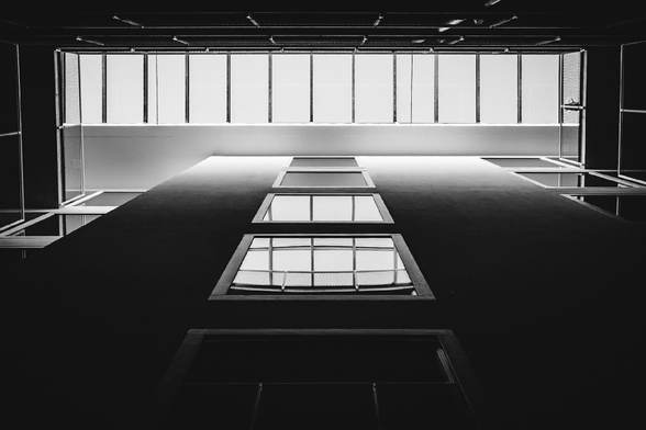 Black and white photo of a modern building’s narrow interior courtyard, viewed from the bottom looking up through a vertical shaft lined with windows. At the top, a grid of frosted glass panels creates a soft glow, contrasting with the dark shadows below.

