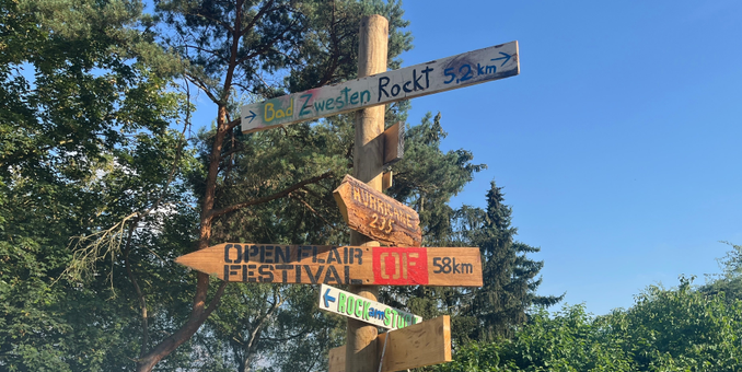 Ein hölzerner Wegweiser vor blauem Himmel und grünem Wald zeigt in verschiedene Richtungen zu bekannten Festivals: „Bad Zwesten Rockt 5,2 km“, „Hurricane 235 km“, „Open Flair Festival 58 km“ und „Rock am Stock“. Die Schilder sind bunt bemalt, teils handbeschriftet und vermitteln eine kreative, DIY-Festivalatmosphäre.