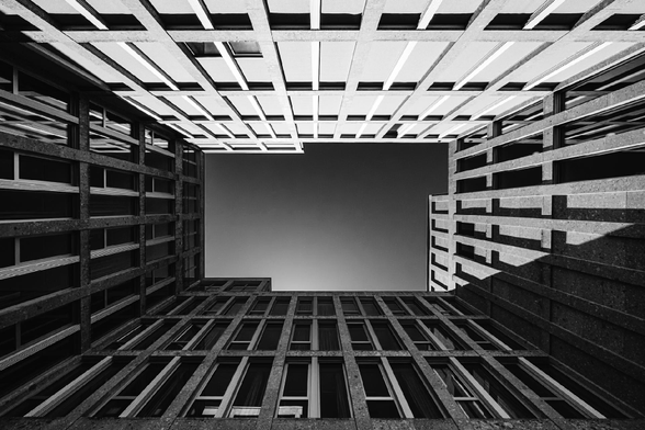 Looking up from a courtyard, the symmetrical walls of a modern building frame the sky in sharp geometric lines. The black and white contrast emphasizes the repeating windows and grid-like patterns of the concrete structure.

