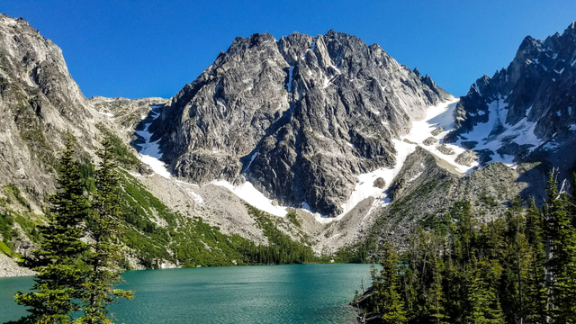 A pristine alpine lake with vibrant turquoise water lies at the base of a towering, jagged granite mountain partially streaked with snow. Dense evergreen trees frame the foreground, while steep rocky slopes rise dramatically to sharp peaks under a clear blue sky.