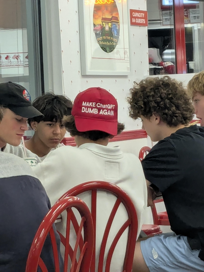 Young men at a table, one with a red ball cap embroidered in white, Make ChatGPT Dumb Again