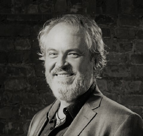 Black and white portrait photo of a smiling, bearded man in a suit, in front of a brick wall. 