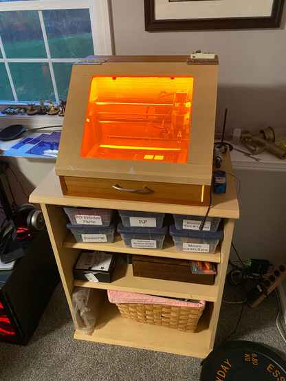 An angled enclosure sits on a shelving unit. The enclosure has a bright orange window with a machine visible inside.