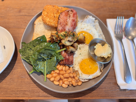 회색 접시에 담긴 풀 잉글리시 브렉퍼스트(full English breakfast). 접시 중앙에는 노른자가 선명한 계란 프라이 2개가 놓여 있고, 그 주변으로 바삭하게 구운 베이컨 2장, 오렌지색 해시브라운 1개, 허브로 양념된 구운 버섯들, 신선한 시금치 잎, 그리고 오렌지색 구운 콩(베이크드 빈즈)이 배치되어 있다. 접시 오른쪽에는 작은 그릇에 담긴 버터가 함께 제공되었다. 식기류로는 포크와 나이프가 접시 오른쪽에 놓여 있으며, 모든 음식이 나무 테이블 위에 정갈하게 세팅되어 있다.