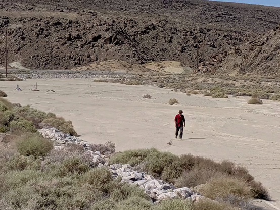 A gif of me in the distance walking along a dry river bed in the desert. I look like I'm skating along and waving at the camera. I'm wearing a brown cowboy hat and a maroon button up shirt
