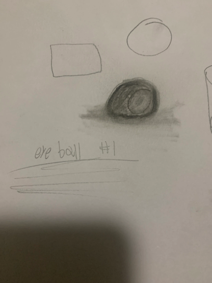 Pencil sketches on white paper that shows basic shapes: a rectangle, circle, shaded oval with shadow, and scribbled text reading "eye ball #1" with lines below.