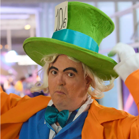 A imagem mostra uma pessoa caracterizada como o Chapeleiro Maluco, um personagem clássico de "Alice no País das Maravilhas." Ele usa um chapéu alto e verde, com uma faixa azul e uma etiqueta que diz "10/6." O figurino inclui um terno colorido, predominantemente laranja, com uma gravata borboleta azul e um ar teatral. A expressão facial é exagerada, destacando sobrancelhas marcadas e um olhar sério ou intrigado, típico de uma performance cômica ou carnavalesca.