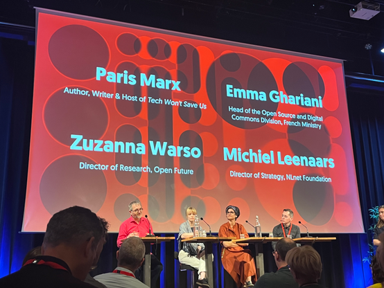 A photo showing four speakers on stage at a conference. From left to right: Michiel Leenaars, Zuzanna Watson, Emma Ghariani and Paris Marx. Above them there’s a tall large screen showing their names