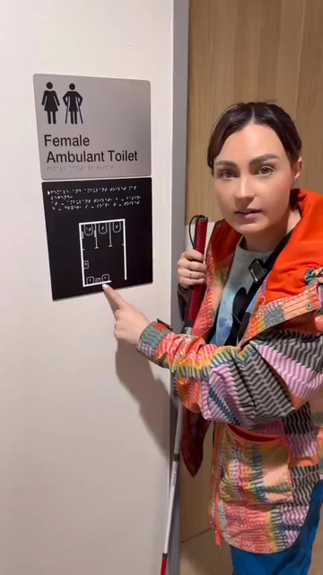 A woman is standing outside a public toilet, holding a white cane and gesturing to signage on the wall which reads "Female Ambulant Toilet". She gestures to the braille sign below which includes a raised, braille labeled diagram of the interior of the washroom. She explains that the braille explains the legend of symbols for the diagram, and how such signage relieves what is often an anxious experience of exploring an unknown washroom without sight, in order to find and use the appropriate facilities.

From @MollyBurkeOfficial on Instagram
