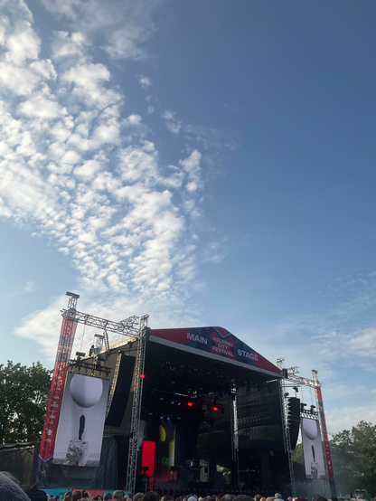 Helsinki outdoors stage with signs HELSINKI CITY FESTIVAL MAIN STAGE, where Jarre is playing in front of an audience. Blue sky with setting, still bright nordic summer sun and a trail of beautiful white clouds.