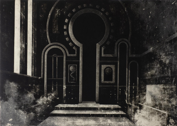 A dimly lit, monochromatic architectural space is depicted in the image. At the center is a large, monumental doorway shaped like an oversized keyhole. The doorway is flanked on either side by symmetrical archways and tall, narrow vertical recesses. The surrounding wall is detailed with ornate framing lines and inset panels, giving a sense of depth and decoration. Two framed motifs resembling abstract medallions or relics are present on each side of the central doorway.

The steps leading up to the entrance are worn and shadowed, creating a dramatic contrast between light and dark areas. The texture throughout the scene is rich, with visible grain and smudging that suggest the surface has been etched, rubbed, or printed. Soft, cloud-like patches spread across the bottom and sides of the composition, adding a slightly mysterious, aged atmosphere.

Overall, the space feels sacred or ceremonial, evoking a timeless, almost religious stillness.