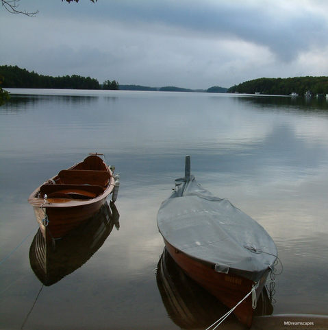 Two disappearing propeller vintage wooden boats moor on the calm water of Lake Joseph, Ontario.  Their shining varnished hulls are mirrored in the still water that shimmers in the twilight. The darkening evening sky and treed islands are in the background.