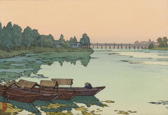 A view of the Yodo River on a sullen, overcast day. In the foreground, three small wooden boats are pulled up by the riverbank; lilypads crowd the shallow water here. A single figure wearing a broad hat is at work in one of the boats, bent over some unseen task. Further up, another little boat is moored among reeds; beyond, trees flank the river as it approaches a bridge downstream, and a few small houses stand beneath the trees closer to the bridge. The river itself is a green colour, while the sky above is dingy and overcast.