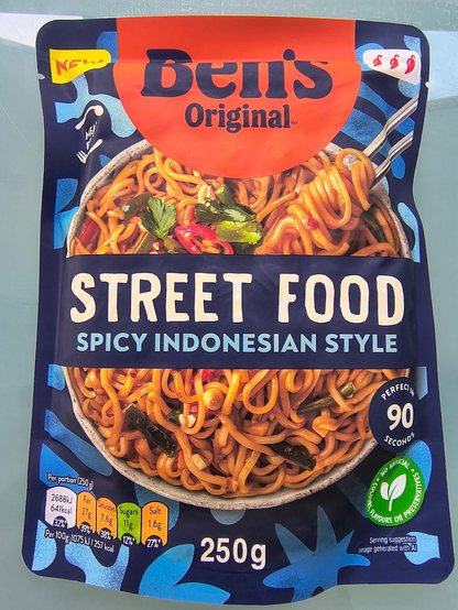 A photograph of a package of Ben's Original Street Food Spicy Indonesian Style noodles, sold in the UK. The package is dominated by a picture of a bowl of noodles, with vegetable-looking things in them. A tiny, tiny note to the bottom-right reads "Serving suggestion. Image generated with AI."