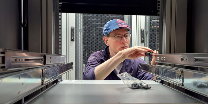 View into some empty space of a 19" computer rack cabinet. A person wearing glasses and a hat with red and blue color is tightening screws on a set of server rails which are being installed in the rack.