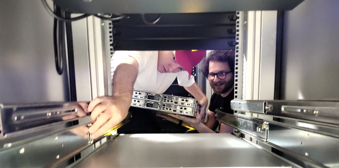 View into empty space of a 19" computer rack cabinet. Two people are preparing to mount a server on freshly installed server rails. The person on the left is wearing a red cap and is using one arm to pull the rail further towards them and the other arm to hold the server. The bearded person on the right is wearing glasses and is holding the server from below with both hands.