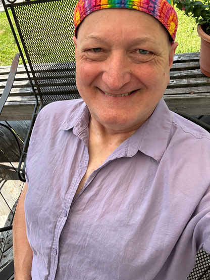 A happy person is outdoors on a deck under a sun umbrella, standing and looking up towards the camera. They are wearing vibrant tye-dye do-rag, a lavender or wisteria blouse (Patagonia calls it “concrete purple”), and a grin a mile wide.