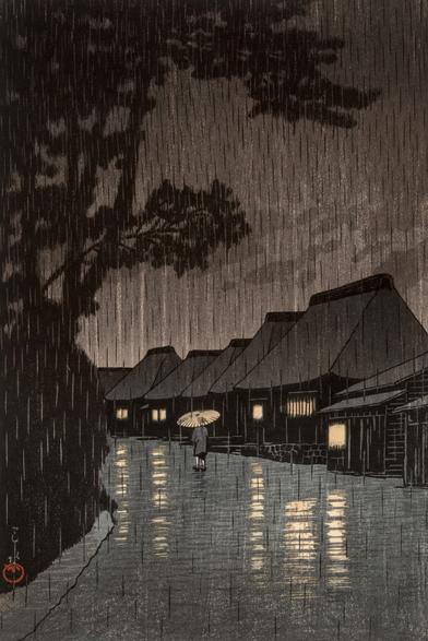 Evening rain falls on a rural street in Maekawa, in modern-day Kanagawa Prefecture. On our left, branches of tall shadowy trees reach out over the road; a row of traditional thatched-roof houses stands on our right, warm lights shining in their windows. The road running down the middle of the image is slick with rain, and steady rain continues to fall from a dim, overcast sky. A solitary pedestrian carries a bamboo umbrella over their head as they walk away from us.