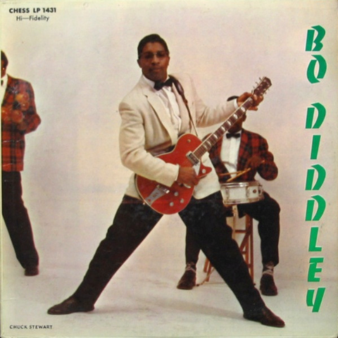 The cover art is a photo of the artist, a black man wearing glasses with tinted lenses, a cream coloured suit jacket, white button up shirt, black bow tie, black pants, and black shoes. He's holding a red guitar and is standing with his legs spread apart, like he's in the middle of a groove. In the background are two band members in matching outfits - red plaid suit jacket, white shirt, black bow tie, black pants, black shoes, their faces not in view. One is sitting on a stool with a drum set up in front of him, drumsticks in hand. The backdrop is white, and the artist's name is written vertically in green letters along the righthand side.