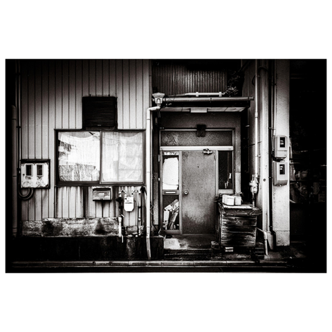 A gritty exterior of what appears to be a back entrance or side façade of a building in Nagoya. The image is rendered in high-contrast black and white, drawing attention to the weathered siding, a rust-streaked window, and a dented metal door with a screen panel. To the right, stacks of wooden boards are topped with a couple of plastic buckets, hinting at some kind of industrious activity—past or present. Pipes, meters, and wires trace their way across the surfaces like urban veins, adding a quiet complexity to the otherwise utilitarian composition.