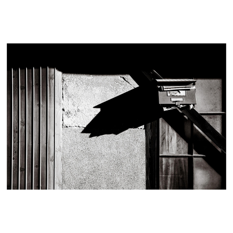 A weathered mailbox juts from a traditional exterior wall, its overstuffed contents creating a dramatic, exaggerated shadow in the harsh afternoon light. The surrounding textures—wood paneling, rough plaster, and frosted glass—form a collage of surfaces slowly surrendering to time.