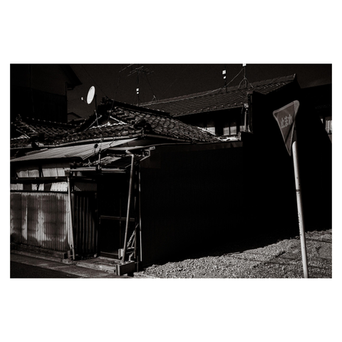 A crooked yield sign leans into frame beside an aging Japanese home. The house, with its layered tile roof and makeshift supports, wears its years unapologetically. A satellite dish sits like a forgotten accessory on a too-old outfit, trying its best to pull the look together. Shadows cut the composition into bold contrasts, emphasizing texture over polish.