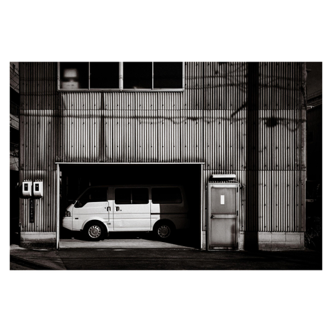 A boxy white van sits snugly inside a corrugated metal garage, half in shadow, half illuminated by the afternoon sun. To its right, an unassuming side door, perhaps leading to an office or workshop, stands quietly, bearing a single sheet of paper—its message unreadable from this distance. Above, shadows from unseen cables and poles crisscross the rippling façade, adding texture to the scene.