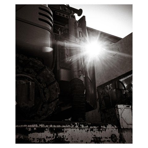A rugged piece of construction equipment sits atop a battered truck bed, its hydraulic arm standing tall against the morning light. The sun bursts through the gaps, casting sharp flares across the frame, but the metal remains unmoved—thick with grime, hardened by labor.