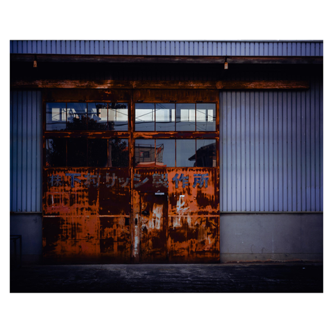 This industrial scene captures a factory's weathered steel door, streaked with rich layers of rust that mimic a painter’s bold strokes. Above, rectangular glass panes reflect the distorted outlines of nearby buildings and utility poles, creating an interplay of texture and light. The corrugated metal walls on either side provide a minimalist frame for the door's rugged, expressive beauty, blending decay and artistry in the heart of the city.