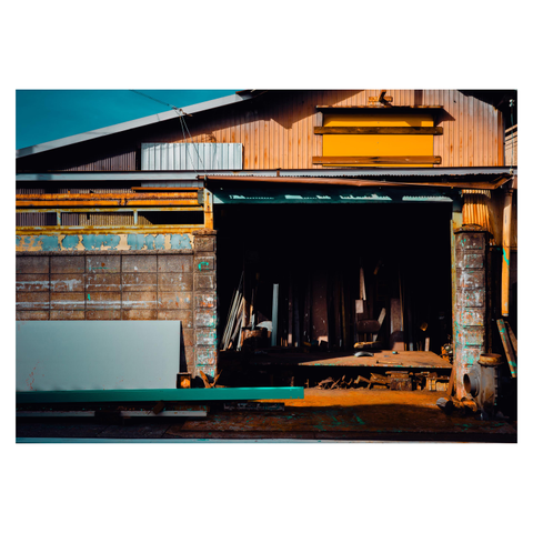 This industrial scene captures the front of an old workshop, its facade a patchwork of rust, faded paint, and sunlight. The dark interior contrasts sharply with the vibrant hues outside, revealing piles of metal and scraps in a state of suspended chaos.
