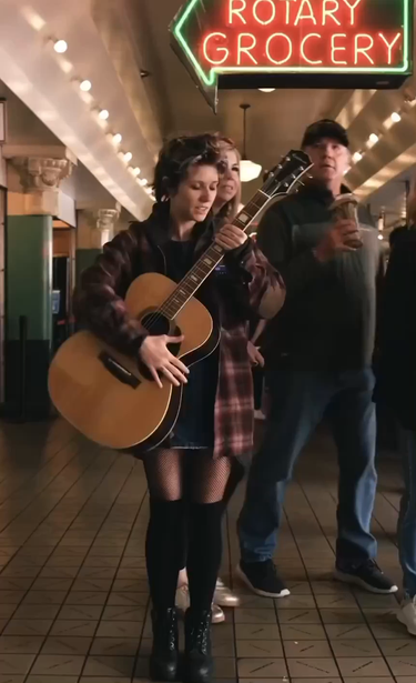 A young woman stands in a crowded marketplace with a guitar and sings a song about eating chips and her distaste for republicans 
