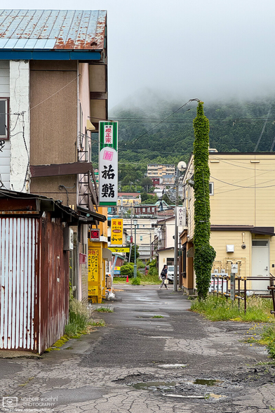 A backstreet alley in the Horaicho district of Hakodate, Japan. Some dated buildings and shop signs in the foreground, and low-hanging clouds over Mount Hakodate in the back.
