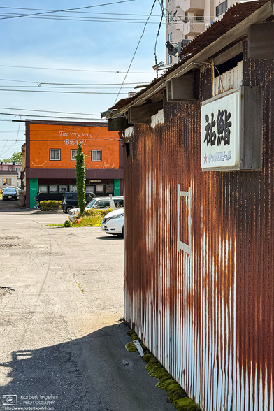 Side street angle in the Horaicho district of Hakodate, Japan. A rusty building with a dated "Sapporo Beer" sign in the foreground, an a restaurant named "The Very Very Beast" in the back.