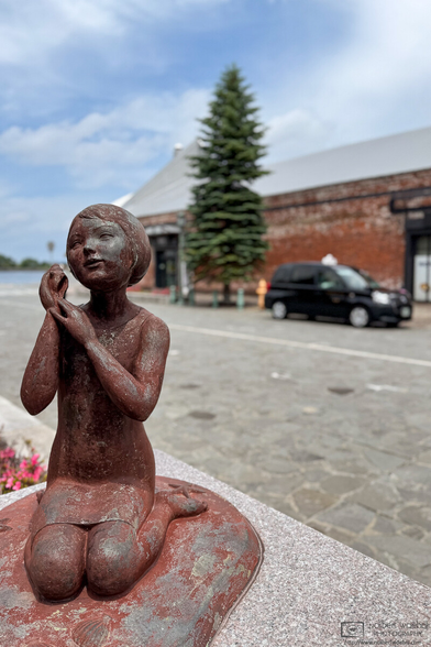 Statue of a girl kneeling on the beach with seashells in the Red Brick Warehouse area of Hakodate in Hokkaido, Japan. In the background, out of focus, a black taxi is waiting outside one of the warehouses.