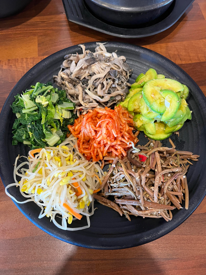 Picture of 5 different colored Namuls on black big plate. 
Namul (Korean: 나물) refers to either a variety of edible greens or leaves or seasoned herbal dishes made of them.