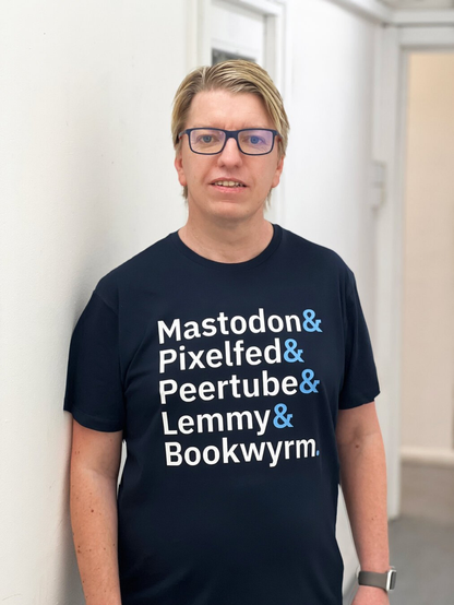 A light skinned man standing in a white corridor. He has blond hair and is wearing glasses. He is wearing a dark navy T-shirt with the words “Mastodon &amp; Pixelfed &amp; Peertube &amp; Lemmy &amp; Bookwyrm.” in bold white text arranged on separate lines.