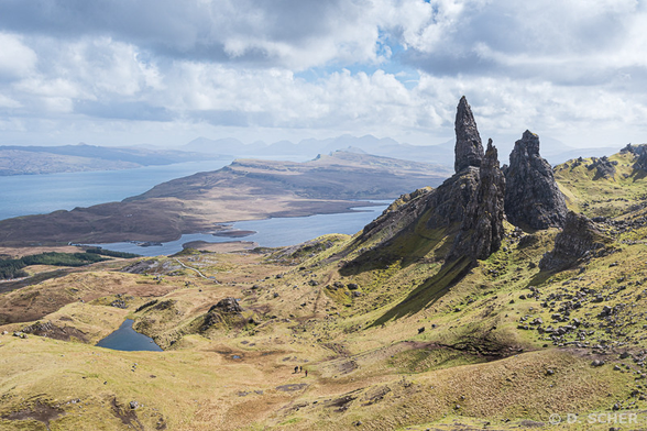 #photography #landscape #mountains #Scotland #Skye

The Old Man of Storr, a 55-meter-high basalt pinnacle, on a sunny day with a few clouds, looking out to sea over an island and then beyond to shores and mountains. 
A small group of hikers can be seen at the bottom of the photo, giving a better idea of the scale.