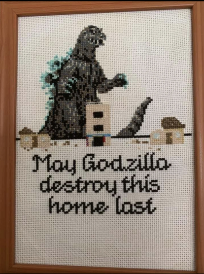 A photo of a needlepoint, cross-stitch in a wooden frame. Godzilla is portrayed walking up to a line of buildings. Under the buildings are the words “May Godzilla destroy this home last.” 
