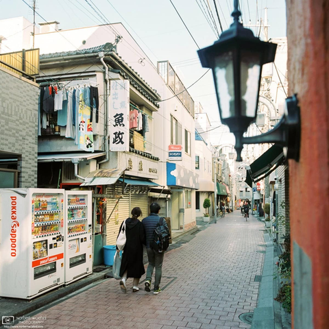Square-format medium-format film photo of a backstreet in Koenji, Tokyo, Japan. Pedestrians are walking on a cobblestone type of pavement. A tight line-up of residential and commercial buildings provides a perspective along the street. In a narrow gap outside an upper-floor window of a building in the foreground, a tight line of laundry is hung out to dry.