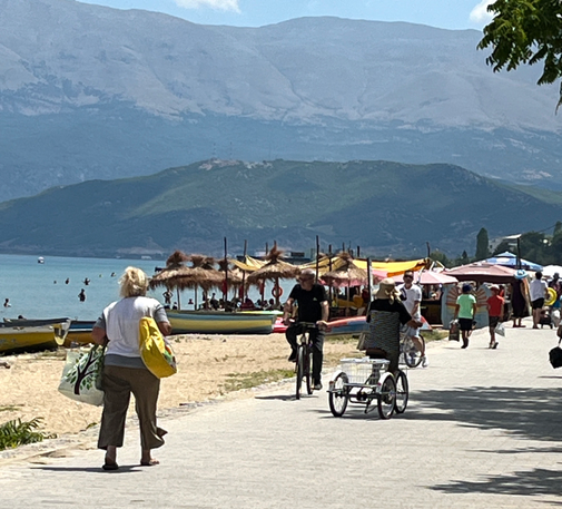 Hot, sunny morning along Lake Ohrid. The mountains form a background to people under straw-covered beach umbrellas. People walk, bike, and trike along the promenade. Including a woman riding away from the camera on an upright delta trike as someone rides towards the camera on an upright bicycle.