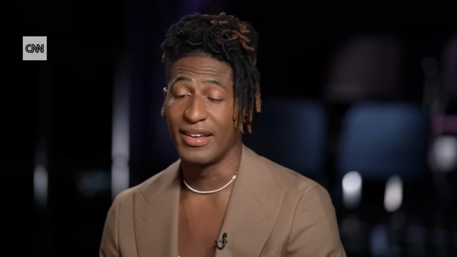 Jon is a good-looking young Black man with with robustly-styled dreadlocks. He is wearing a tan suit coat, partially open over a low-cut white t-shirt. He’s sitting at a piano in what appears to be a small studio setting. Chris is a middle-aged white man with simple glasses. He’s wearing a suit with a bright-red pocket square over a solid-blue dress shirt and a striped tie.