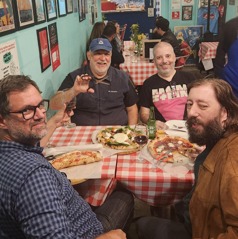 Quatro homens estão sentados em uma mesa de pizzaria com toalha xadrez vermelha e branca, sorrindo e brindando com taças de vinho tinto e garrafas de cerveja. Há três pizzas grandes sobre a mesa, cada uma com coberturas variadas. Ao fundo, é possível ver outras mesas ocupadas, pôsteres decorativos nas paredes azul-claro e um ambiente descontraído. À esquerda está Érico Andrei, segurando uma taça de vinho e olhando para a câmera. O homem ao lado dele, Luciano Ramalho,usa boné azul e faz um gesto com a mão enquanto sorri. À direita, dois outros homens, Osvaldo Santana e Alexandre Villares, também sorriem, um deles vestindo uma camiseta preta com estampa rosa e outro com casaco marrom e barba longa. A atmosfera da imagem é de confraternização entre amigos em um restaurante acolhedor.

