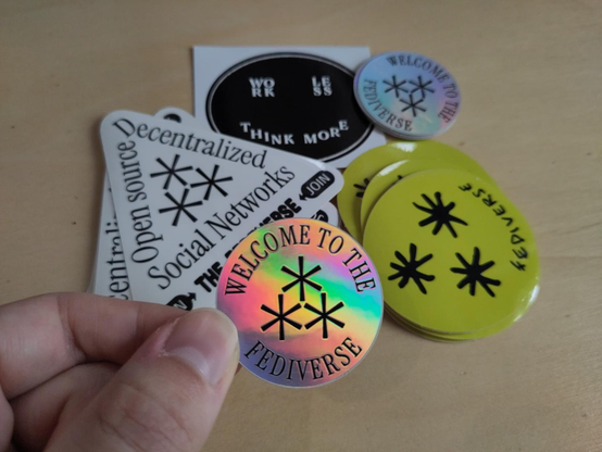 Pack of stickers designed by Raphaël Bastide: 3 Fediverse stickers + 1 Work Les Think More.
I'm holding a small silver circle with iridescent highlights, which bas the words “Welcome to the Fediverse” around the Fediverse symbol ⁂