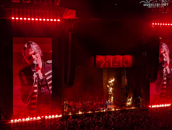 My Chemical Romance performing on stage under a stylized MCR logo with lots of red lights and pyrotechnics around them. Gerard Way is visible singing on two massive screens flanking the stage.