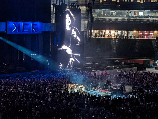 A cellist performing on a b-stage way out in front of the main stage, in the midst of a huge crowd of people. They are dramatically back-lit with a spotlight coming from above the main stage, below the stylized MCR logo. They're visible in dramatic black and white on the massive screen next to the stage.