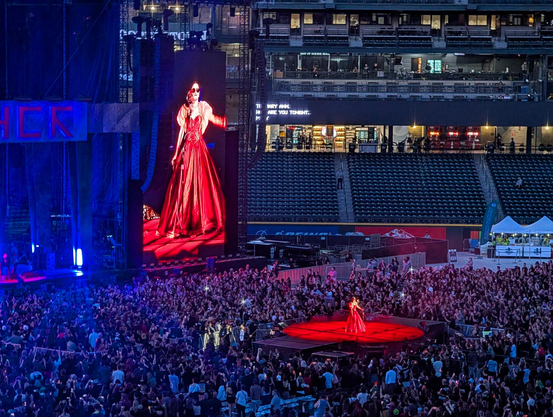 A performer in an intricate red dress wearing round dark glasses, singing on a b-stage way out in front of the main stage in the midst of the crowd. She is bathed in a perfect circle of red light, and visible more clearly on the massive screen next to the main stage.
