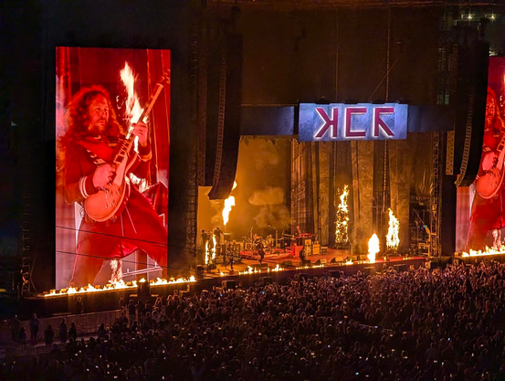 My Chemical Romance performing on stage with gratuitous amounts of pyrotechnic fire, below a stylized MCR logo. Guitarist Ray Toro is visible shredding on the massive screens flanking the stage.
