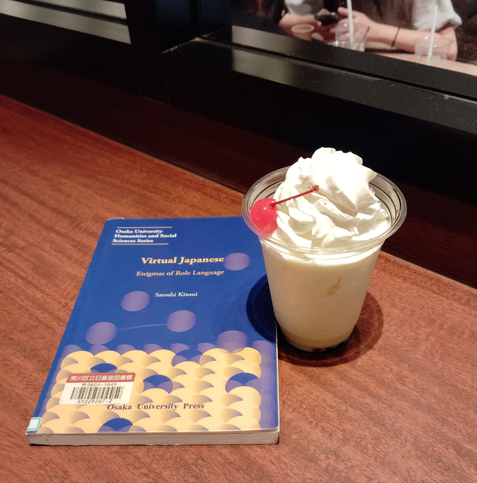 Photo is of a wooden counter. The blue paperback library book is on the counter and a green shake with whipped cream and maraschino cherry in a plastic cup to the right. A woman's arms using her smartphone can be seen through the window but you can't see her face