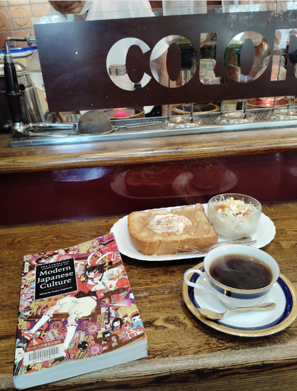 Photo is of a wooden counter. On it is the paperback library book with an illustration from manga with a Japanese girl floating amongst Japanese dolls. To the right is a mug of black coffee. The white mug and saucer have a gold and blue ring. Behind it is thick cinnamon toast with white butter and a small salad in a clear small bowl. Above you can see a glass partition with a thick black rectangle with "COLOR" OF "Colorado" and siphon coffee makers behind it along with the elderly Japanese barista whose face cannot be seen