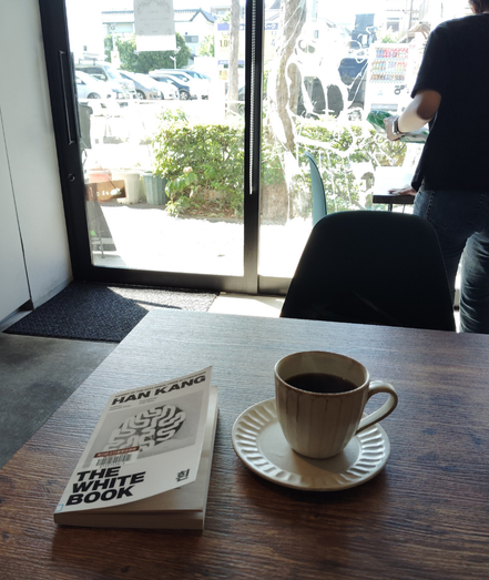 Photo is of inside a cafe. On a dark wooden table is a white mig & edge fluted saucer of black coffee. To the left is the white paperback library book with white bending artwork that looks kinda like a brain. You can see the sidewalk through the brightly lit window. Before the window you can see the partial back of the woman barista cleaning a window side table 